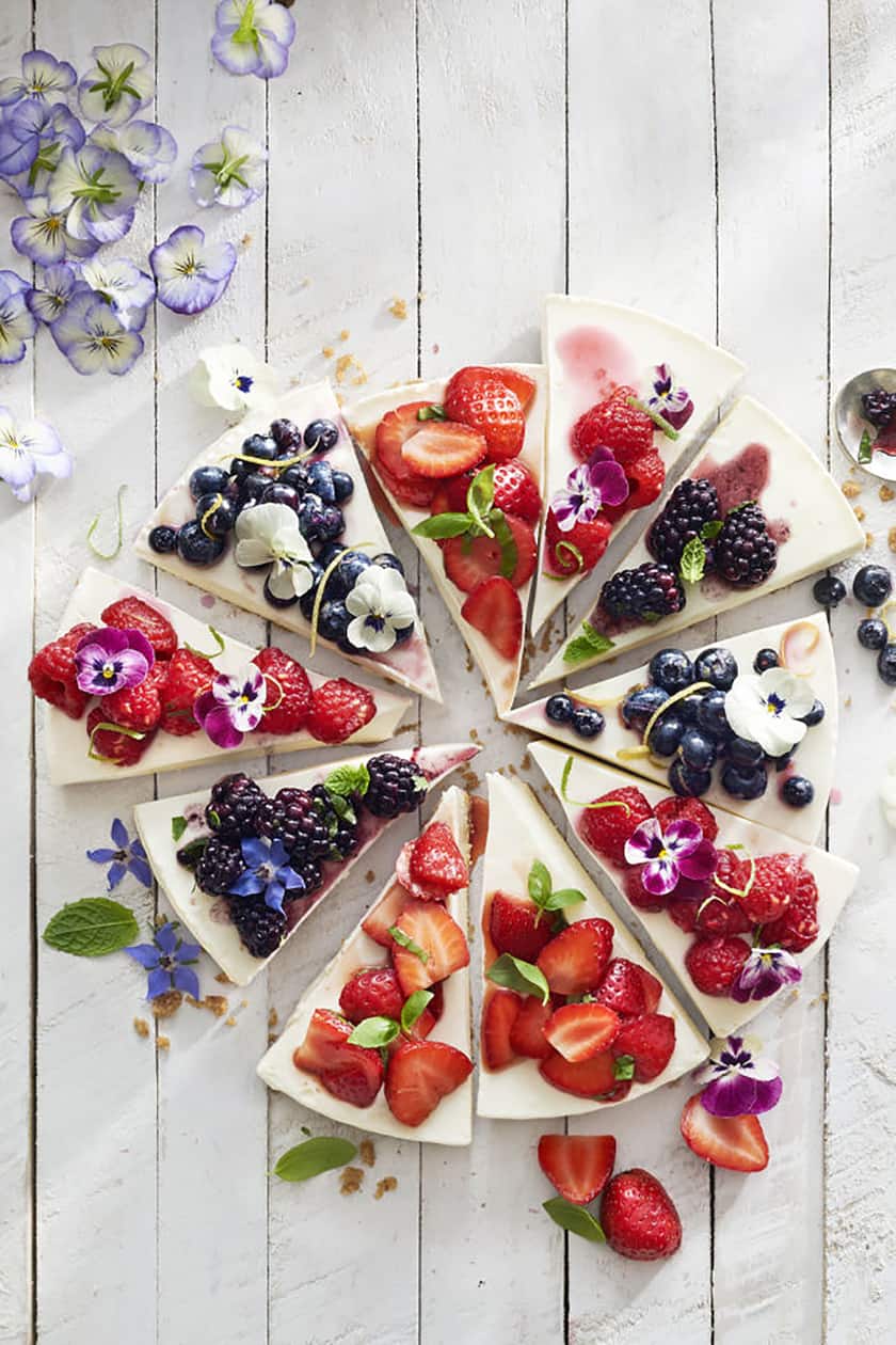 A round cheesecake, perfect for a Mother's Day Breakfast, sliced into pieces and topped with assorted fresh berries and edible flowers, is arranged on a white wooden surface.