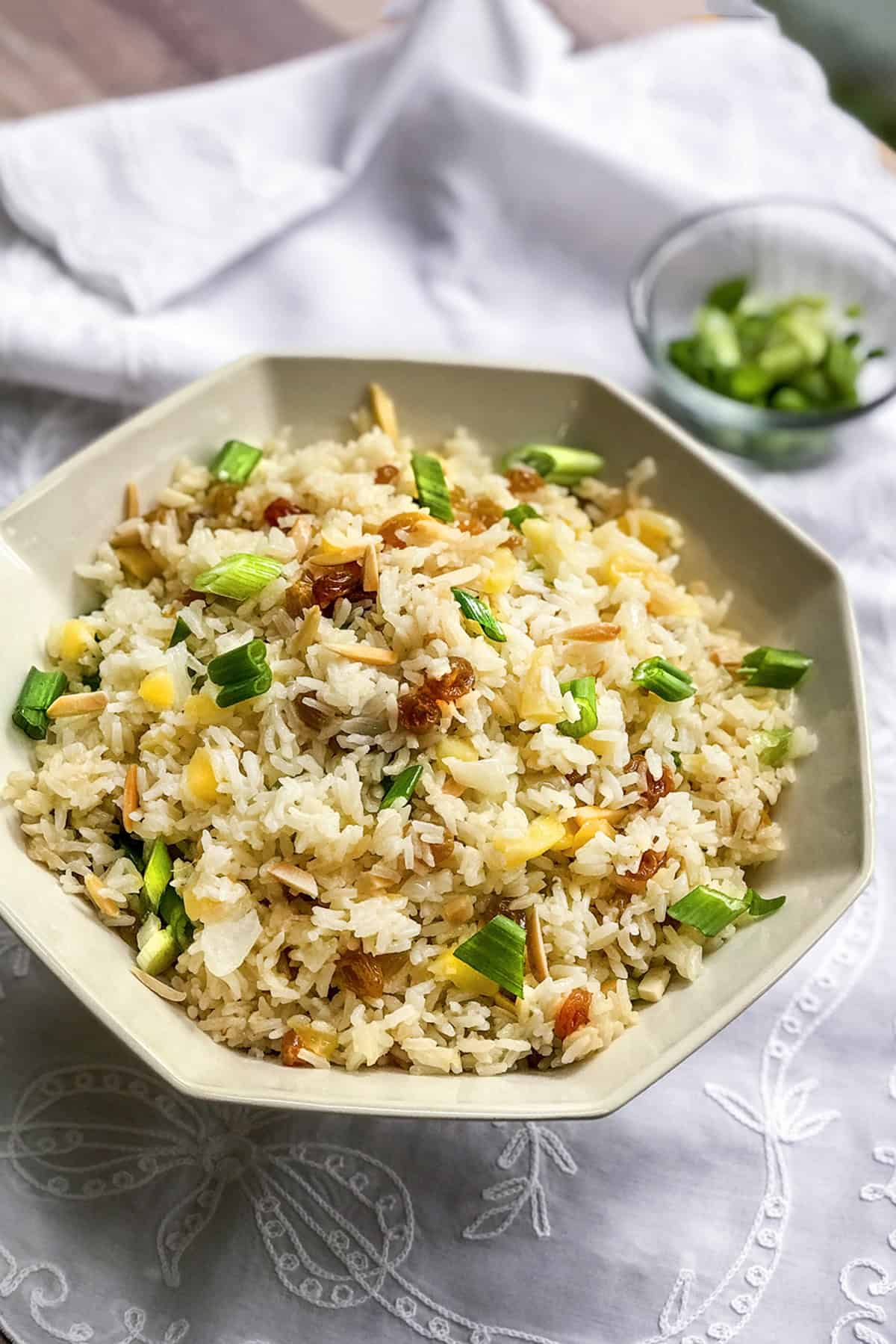 A bowl of rice pilaf fried rice garnished with chopped green onions, pineapple chunks, and slivered almonds, placed on a white tablecloth.