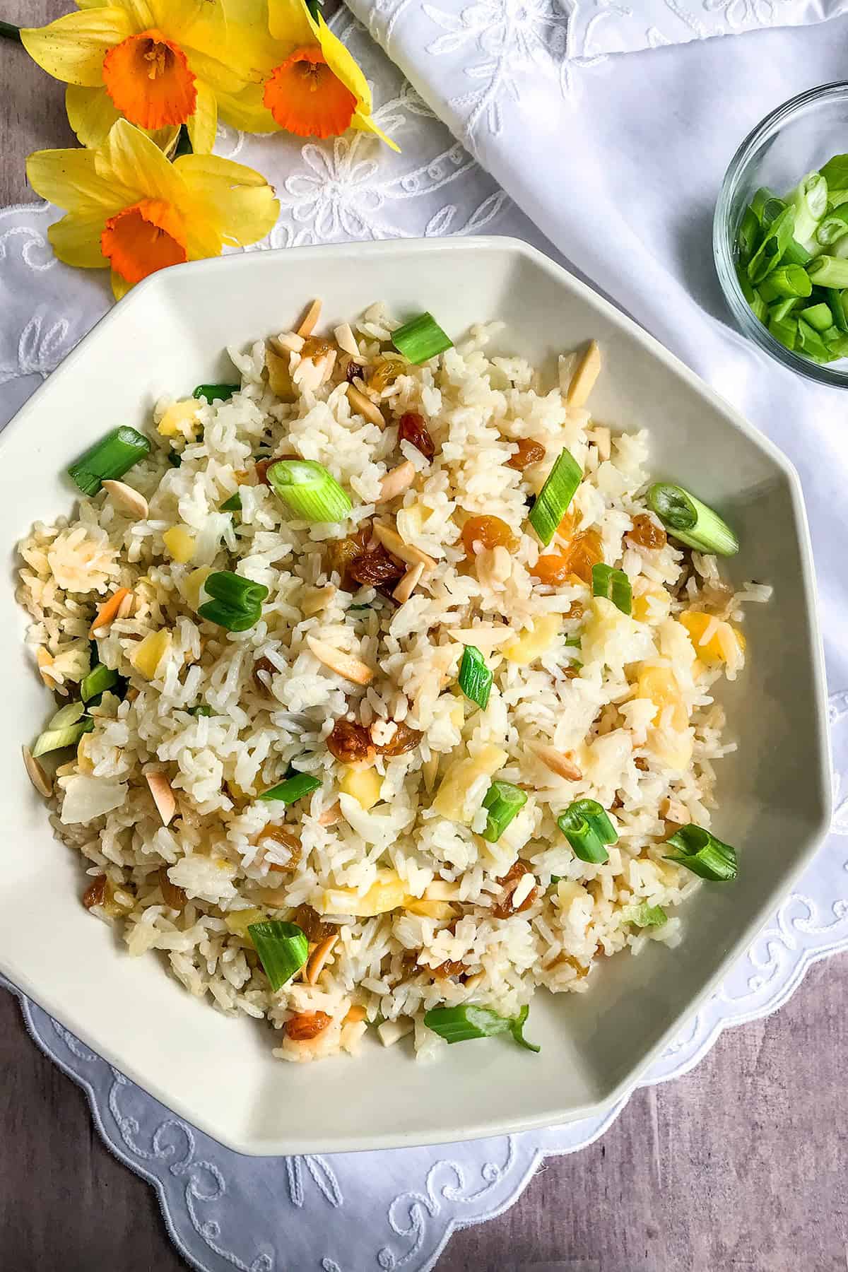 A bowl of rice pilaf fried rice garnished with chopped green onions, raisins, and slivered almonds, placed on a white cloth next to yellow flowers and a small bowl of green onions.