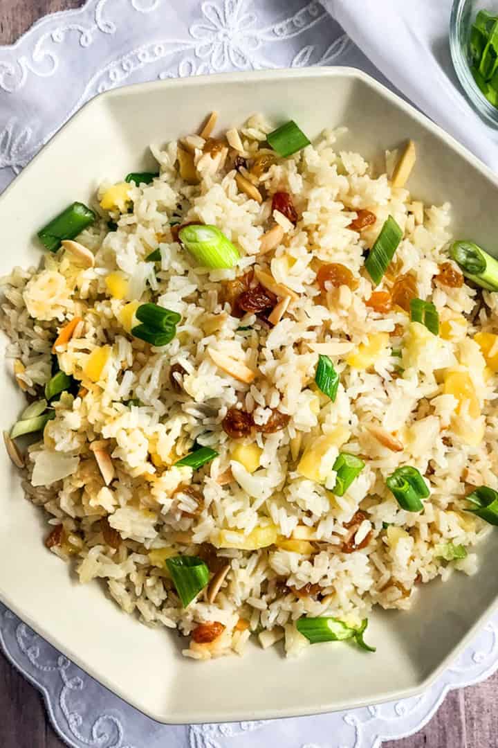 A bowl of rice pilaf garnished with chopped green onions, raisins, and slivered almonds, placed on a white cloth with a small bowl of green onions nearby.