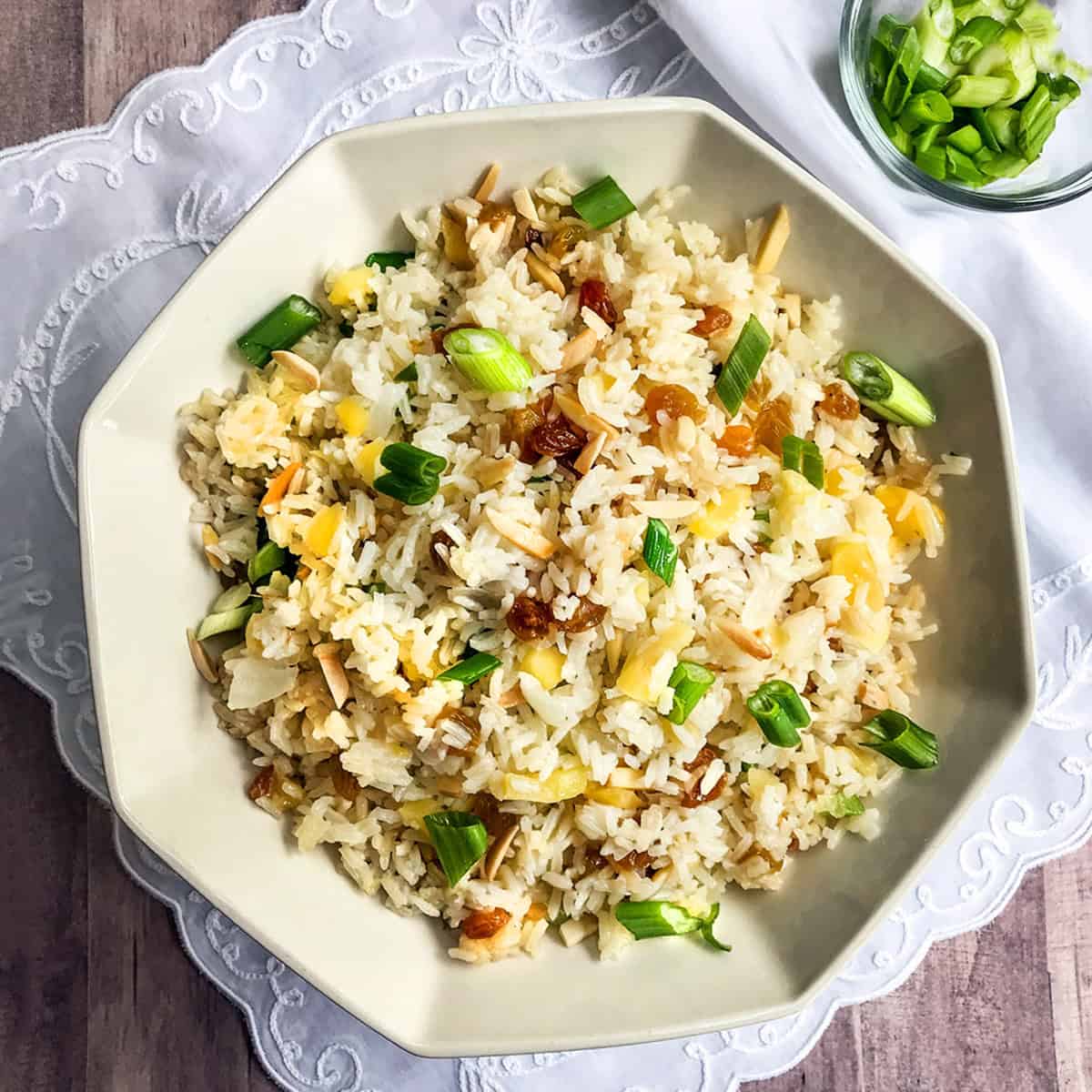 A bowl of rice pilaf garnished with chopped green onions, raisins, and slivered almonds, placed on a white cloth with a small bowl of green onions nearby.