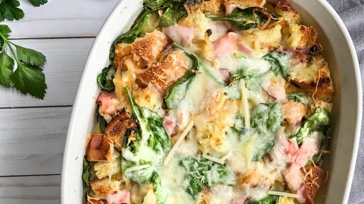 A savory breakfast strata with spinach, ham, cheese, and bread cubes is baked to perfection in a white dish, set on a light wooden table with plates, pepper, and grated cheese nearby.