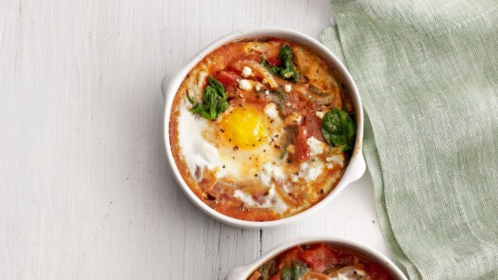 Three small white bowls with baked eggs, tomatoes, spinach, and cheese make a perfect Mother's Day breakfast, served on a white surface beside two silver spoons, a green cloth, and a small dish of black pepper.