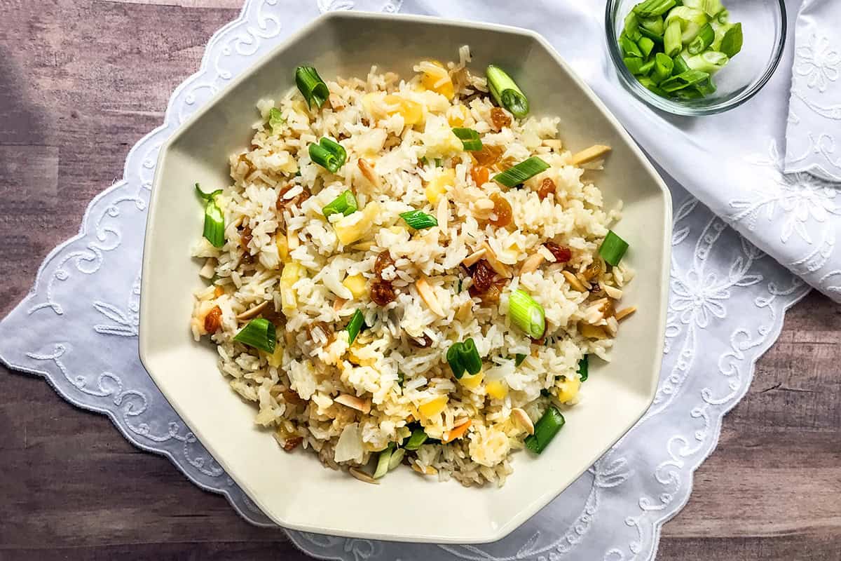 A white plate of rice pilaf fried with chopped green onions, almonds, and raisins sits on a white embroidered napkin, accompanied by a small bowl of green onions nearby.