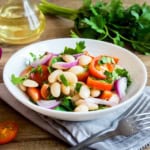 A white bowl filled with a White Bean Olive Salad featuring tomato slices, red onion, and fresh parsley sits on a wooden table with a fork beside it.