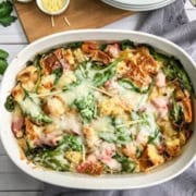 A savory strata casserole filled with bread pieces, spinach, ham, and melted cheese sits on a gray napkin next to a wooden board and fresh parsley.