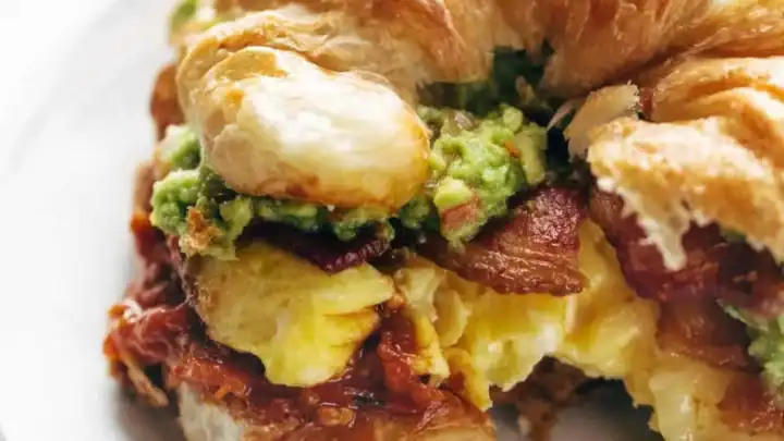 A croissant sandwich filled with scrambled eggs, bacon, avocado, and possibly tomato or salsa, served on a white plate—one of the easy Mother’s Day breakfast recipes that makes mornings extra special.