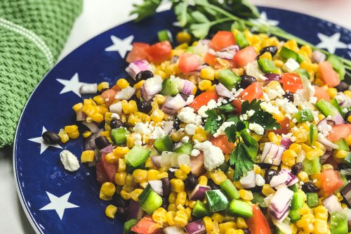 A blue plate with white stars holds a vibrant Mexican corn salad with diced tomatoes, cucumbers, red onions, black beans, and crumbled cheese. A green napkin and vine tomatoes are in the background.