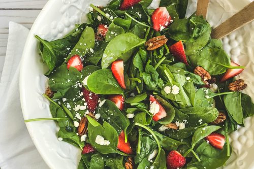 A white plate of strawberry spinach salad with pecans and crumbled cheese sits on a white napkin. Wooden serving utensils rest in the salad, and a small jar of dressing is nearby on a white wooden surface.