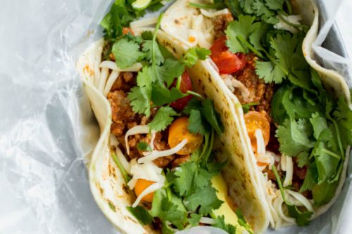 Two soft tacos filled with ground meat, shredded cheese, cherry tomatoes, and fresh cilantro-perfect for summer-garnished with lime wedges and served on parchment paper. Try this Summer Slow Cooker favorite for an easy meal!.
