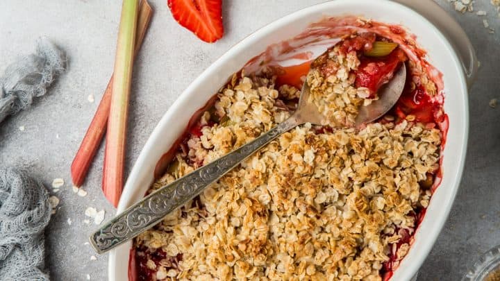 A freshly baked strawberry rhubarb crumble sits in a white dish with a serving spoon, surrounded by fresh strawberries, rhubarb, and a jug of cream.
