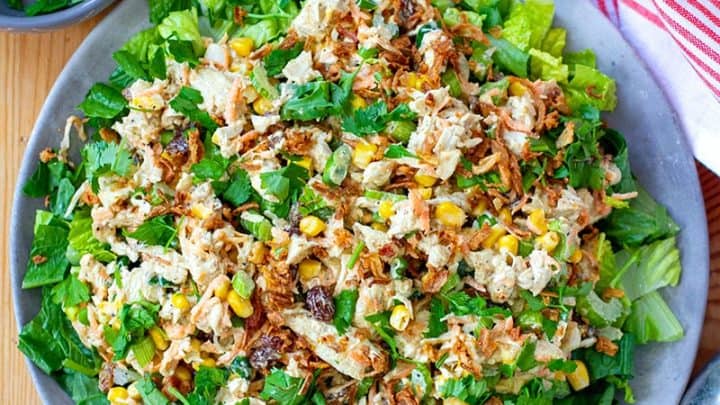 A plate of chopped salad with shredded chicken, greens, corn, green onions, and fried onions—perfect for summer instant pot recipes—surrounded by small bowls of herbs, fried onions, a spice jar, and a red-striped towel.