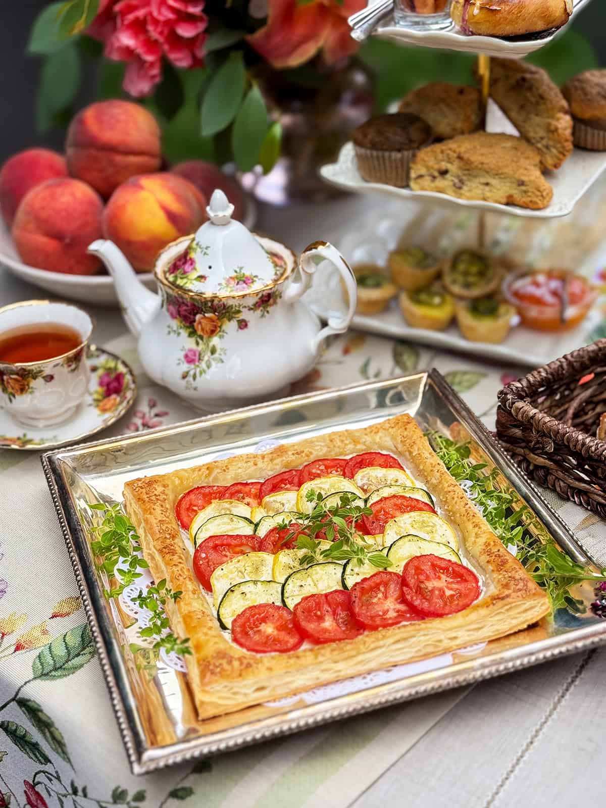 A square puff pastry tart inspired by Tian Provençal, topped with tomato and zucchini slices, rests on a silver tray set atop a floral tablecloth, accompanied by tea, peaches, and assorted pastries in the background.