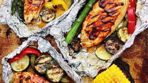 Grilled chicken breasts with vegetables in foil packets, served alongside grilled corn on the cob on a rustic baking sheet—perfect for fans of easy foil packet recipes.