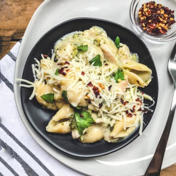 A black plate of instant pot mac and cheese shells in creamy sauce, topped with shredded cheese, red pepper flakes, and herbs, sits on a white plate with a spoon and a small bowl of red pepper flakes nearby.