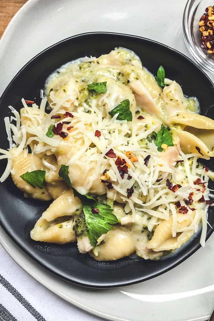A black plate of instant pot mac and cheese shells in creamy sauce, topped with shredded cheese, red pepper flakes, and herbs, sits on a white plate with a spoon and a small bowl of red pepper flakes nearby.