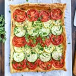 Square tart inspired by Tian Provençal, topped with alternating rows of sliced tomatoes, zucchini, and yellow squash, garnished with chopped herbs, on a parchment-lined surface.