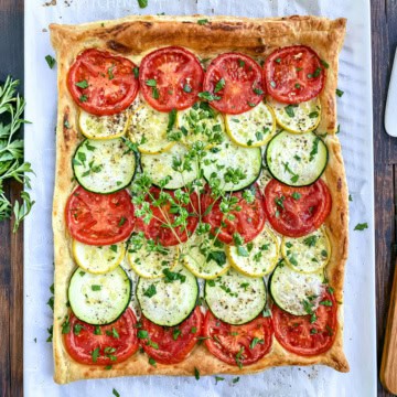 Square tart inspired by Tian Provençal, topped with alternating rows of sliced tomatoes, zucchini, and yellow squash, garnished with chopped herbs, on a parchment-lined surface.