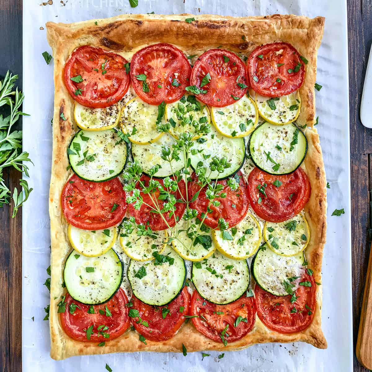 Square tart inspired by Tian Provençal, topped with alternating rows of sliced tomatoes, zucchini, and yellow squash, garnished with chopped herbs, on a parchment-lined surface.