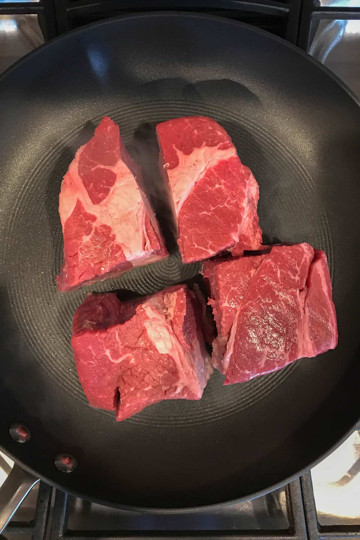 Four raw beef chunks are evenly spaced in a black skillet on a stovetop, ready to be cooked for Mexican Shredded Beef Tacos. The marbling and texture of the red meat stand out against the pan’s surface.