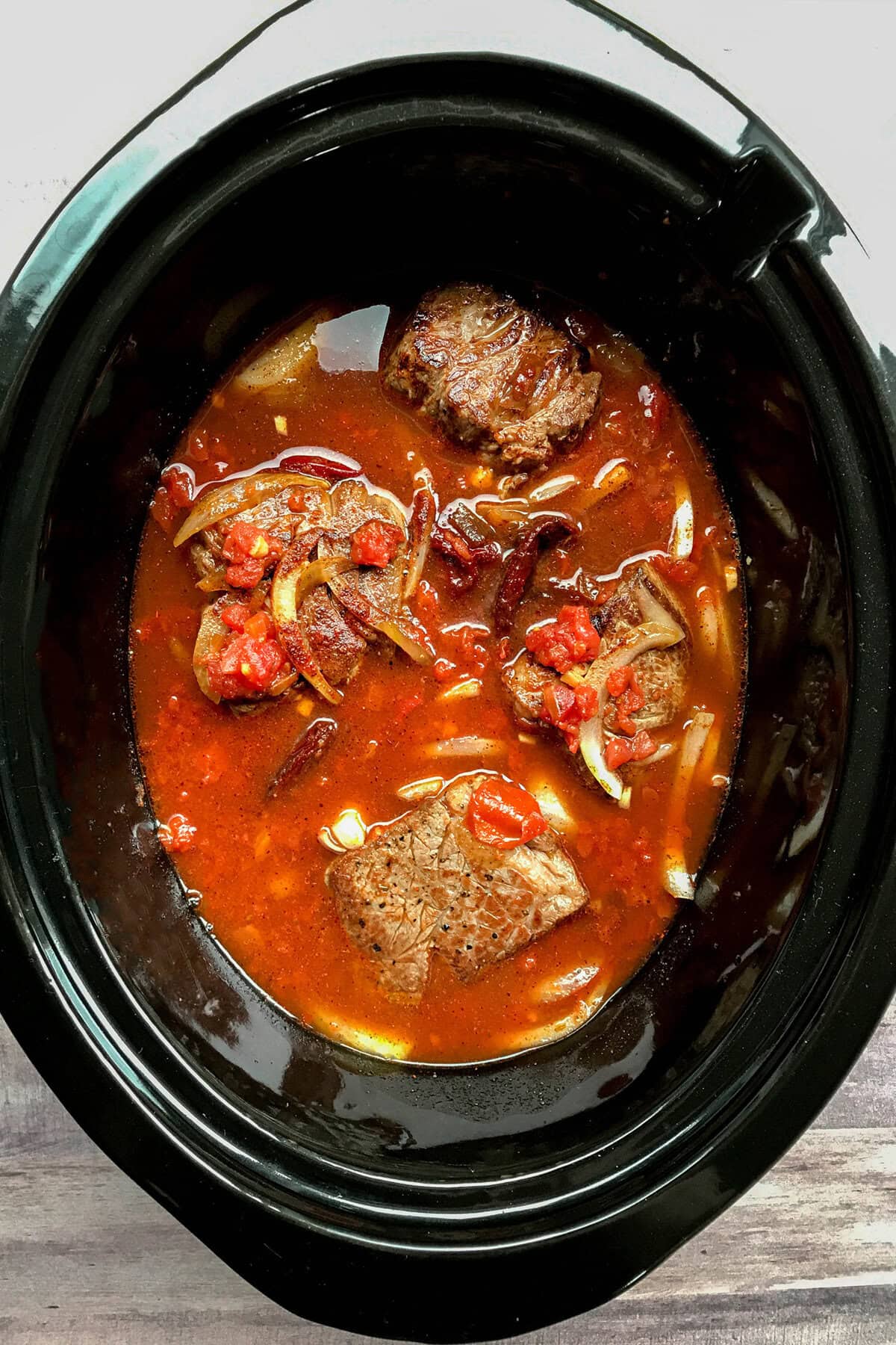 A slow cooker filled with chunks of beef, sliced onions, tomatoes, and a rich red broth—perfect for creating flavorful Mexican Shredded Beef Tacos. The ingredients are partially submerged in the liquid, ready for slow cooking.