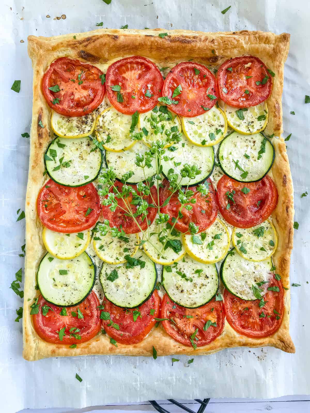 A Tian Provençal puff pastry tart, topped with alternating rows of sliced tomatoes, green and yellow zucchini, and sprinkled with herbs on a sheet of parchment paper.