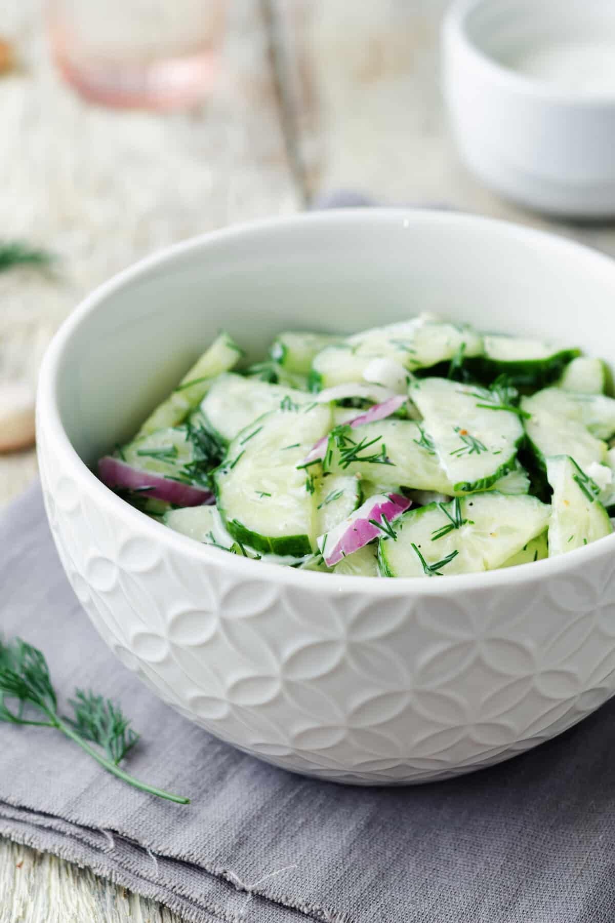 A white bowl filled with cucumber salad, crisp red onion slices, and fresh dill sits on a gray cloth napkin.