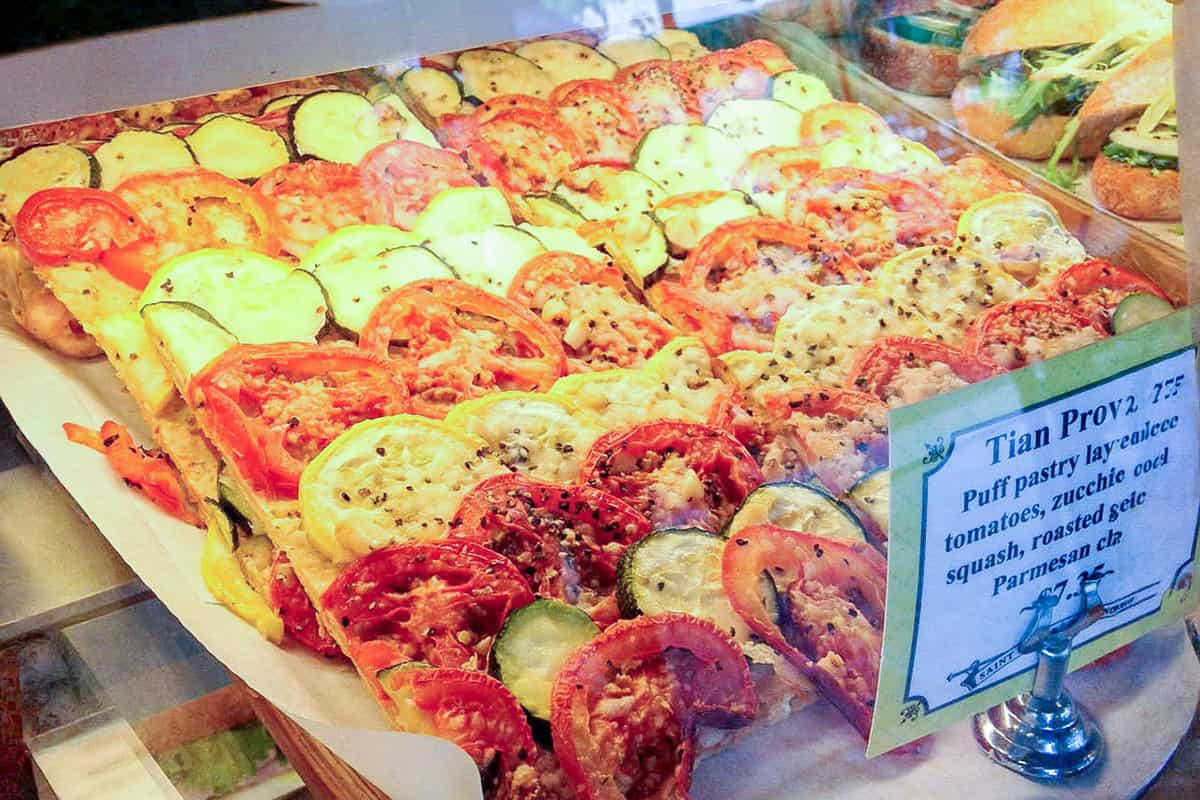 Rectangular puff pastry tart inspired by Tian Provençal, topped with rows of sliced tomatoes, yellow squash, and zucchini, displayed in a bakery case with a sign describing the ingredients.