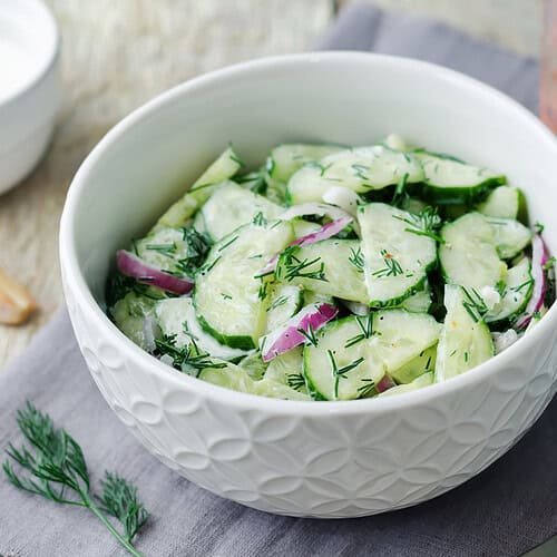 A white bowl filled with cucumber salad mixed with sliced red onions and dill sits on a napkin, accompanied by a glass of water and a small bowl of dressing nearby.