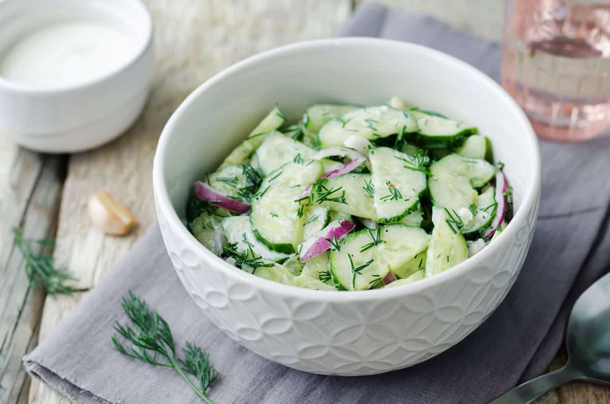 A white bowl filled with cucumber salad mixed with sliced red onions and dill sits on a napkin, accompanied by a glass of water and a small bowl of dressing nearby.