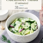 A white bowl of creamy cucumber salad with dill and red onion sits on a gray napkin; text above reads "Easy Creamy Cucumber Salad (5 minutes) 31Daily.com".