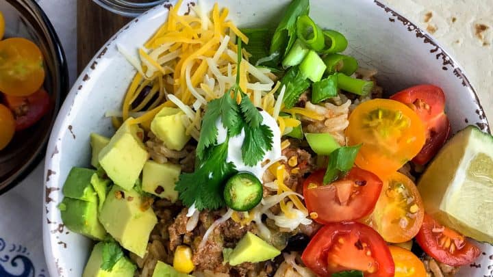 A turkey burrito bowl featuring shredded turkey, rice, avocado, cheese, tomatoes, lettuce, corn, and sour cream, topped with jalapeños and lime, served with flatbread on the side.