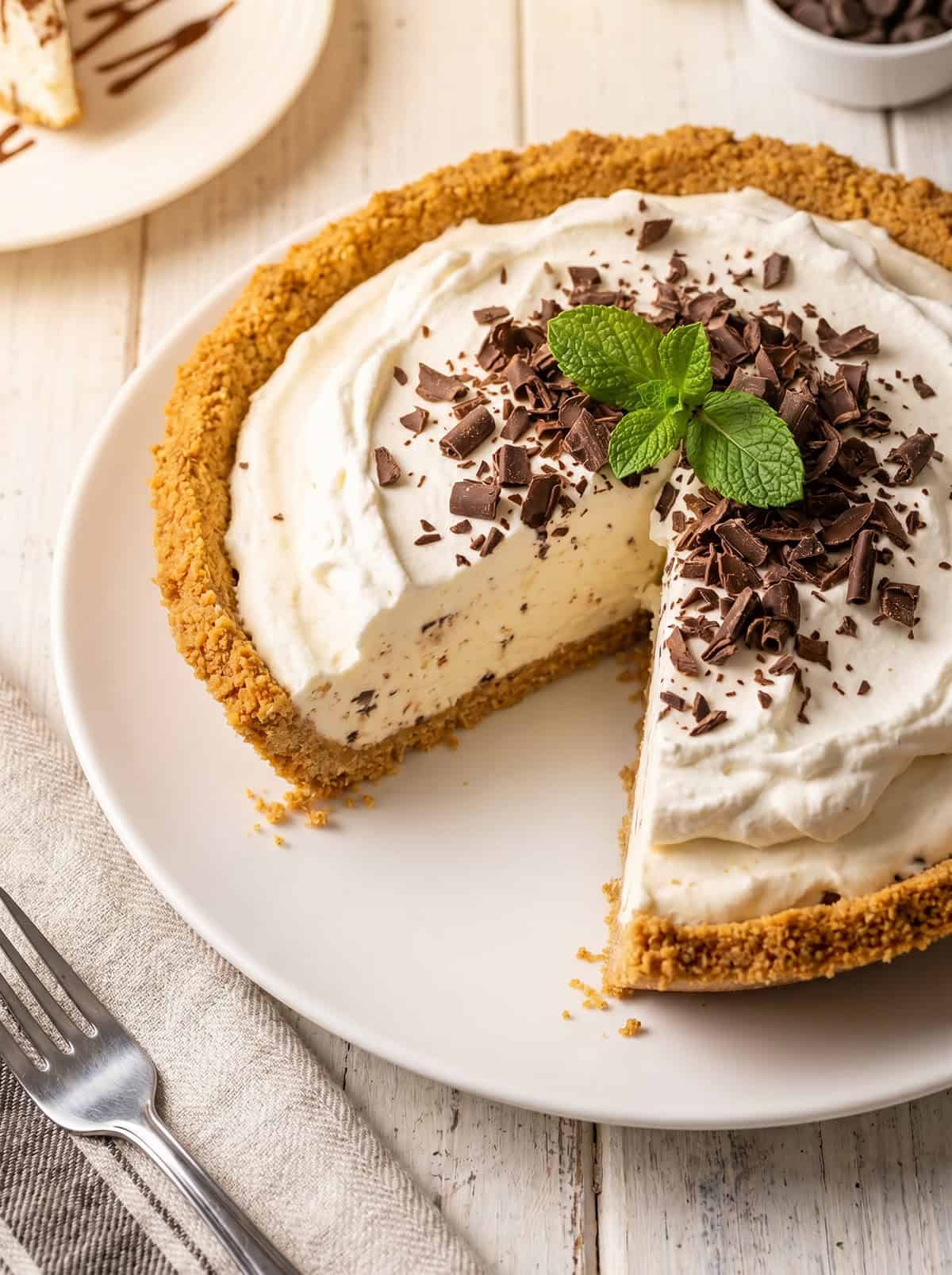 A decadent Ice Cream Pie with a graham cracker crust, topped with whipped cream, chocolate shavings, and fresh mint leaves. One slice is missing. The pie sits on a white plate on a wooden table.