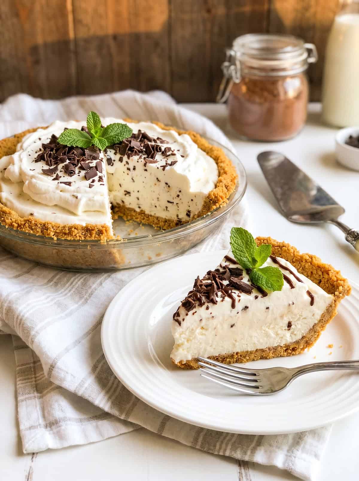 A slice of decadent ice cream pie with chocolate shavings and mint garnish sits on a plate, with the rest of the pie in a glass dish behind it.