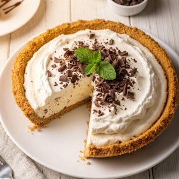 A decadent Ice Cream Pie with a graham cracker crust is topped with whipped cream, chocolate shavings, and mint leaves. One slice has been removed.
