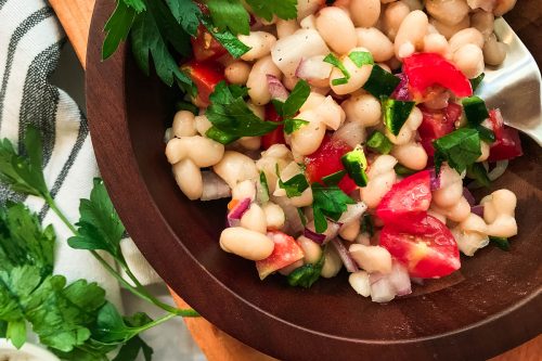 A wooden bowl filled with White Bean Salad, cherry tomatoes, red onion, parsley, and green peppers sits on a wooden board, with a spoon inside and fresh herbs nearby.