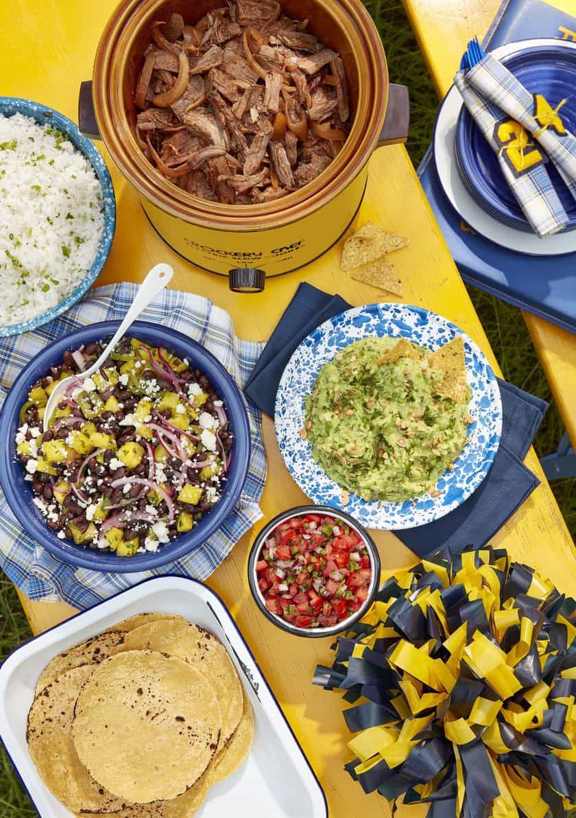 Overhead view of a table with shredded meat in a slow cooker, white rice, bean salad, guacamole, salsa, tortillas, plates, and a blue and yellow pom-pom-the perfect spread for Game Day Recipes.