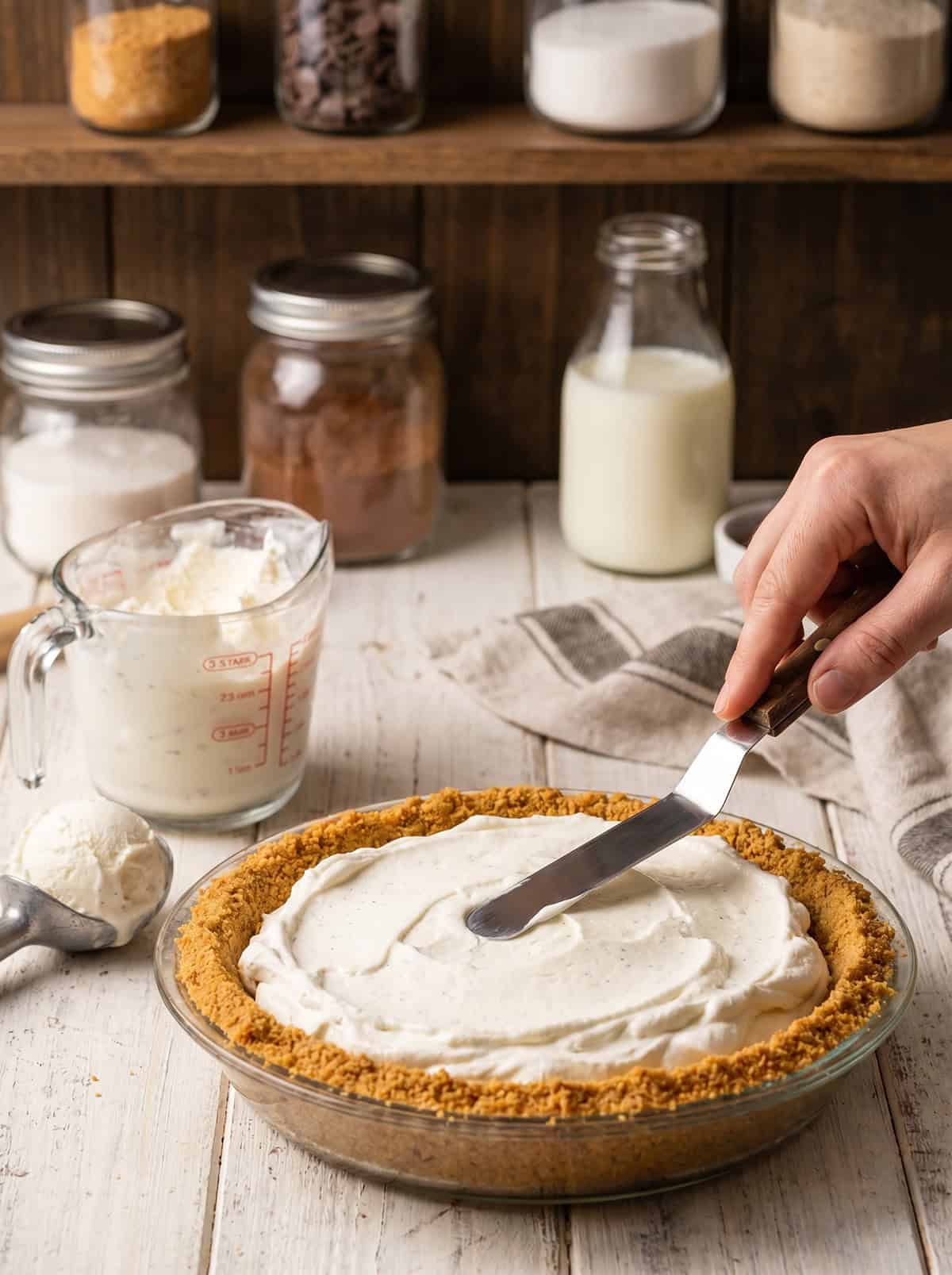A hand spreads cream filling onto a pie crust with a spatula, preparing the base for a delicious Ice Cream Pie. Ingredients such as milk, flour, and cocoa are visible on shelves in the background.