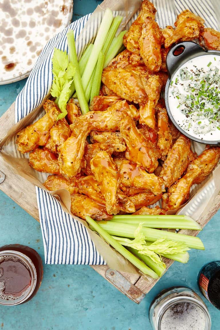 A tray of buffalo chicken wings with celery sticks and ranch dip sits on a blue table next to drinks and an empty plate-perfect for your Game Day Recipes lineup.