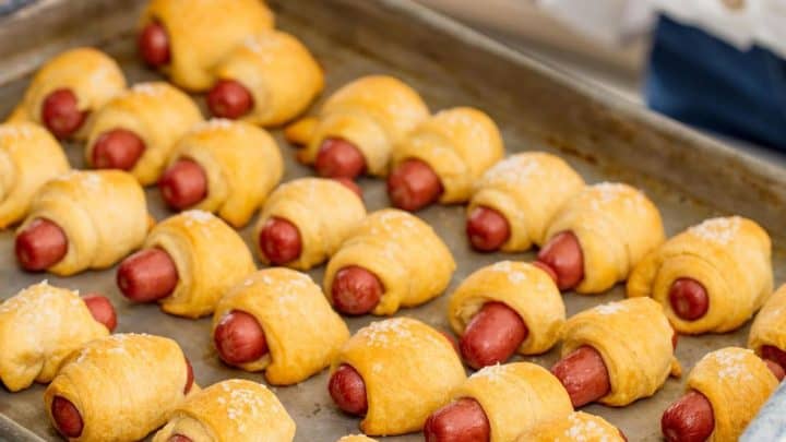 A person holds a baking tray filled with freshly baked pigs in a blanket, made of mini sausages wrapped in golden crescent roll dough—perfect Game Day Appetizers for any crowd.