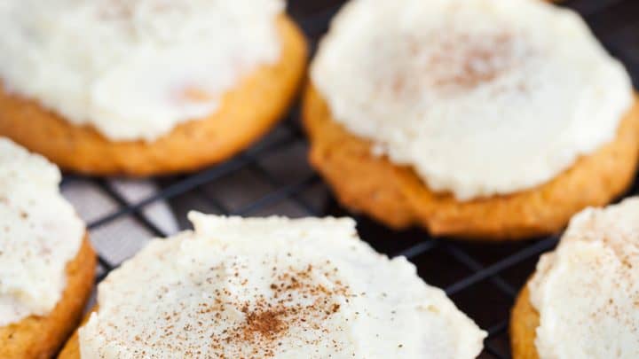 Frosted Pumpkin Spice Cookies with white frosting and a sprinkle of cinnamon are cooling on a black wire rack.