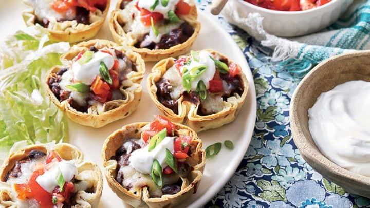 A plate of mini taco cups topped with cheese, diced tomatoes, and green onions makes the perfect Game Day Appetizer, served with shredded lettuce, salsa, sliced green onions, and sour cream on a patterned tablecloth.