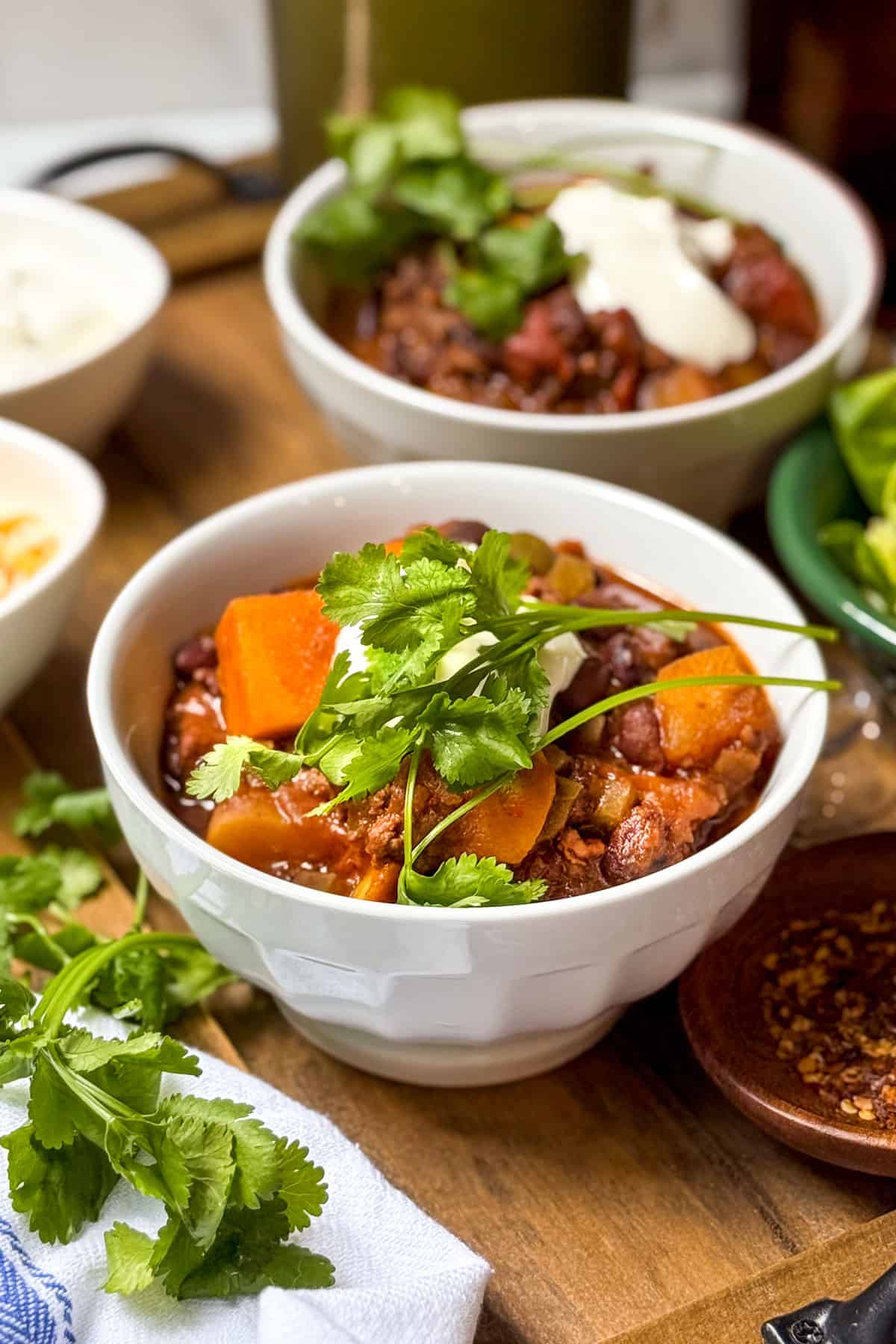 Two bowls of beef and butternut squash chili in white bowls and topped with fresh cilantro, sour cream, and shredded cheese.