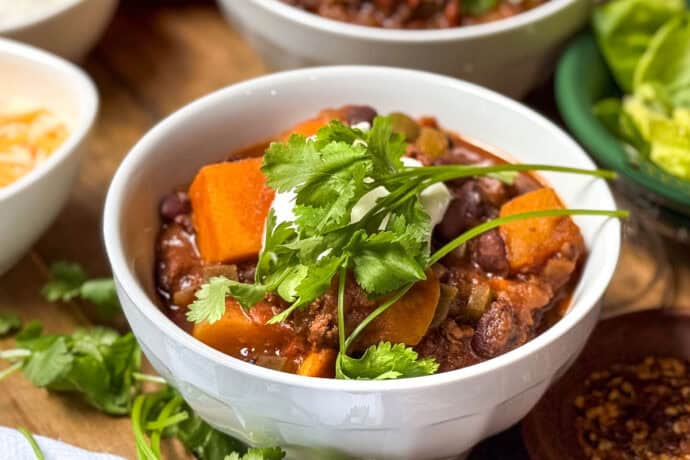 Bowl of Beef and Butternut Squash Chili, garnished with fresh cilantro, in a bowl with shredded cheese and sour cream on the side.