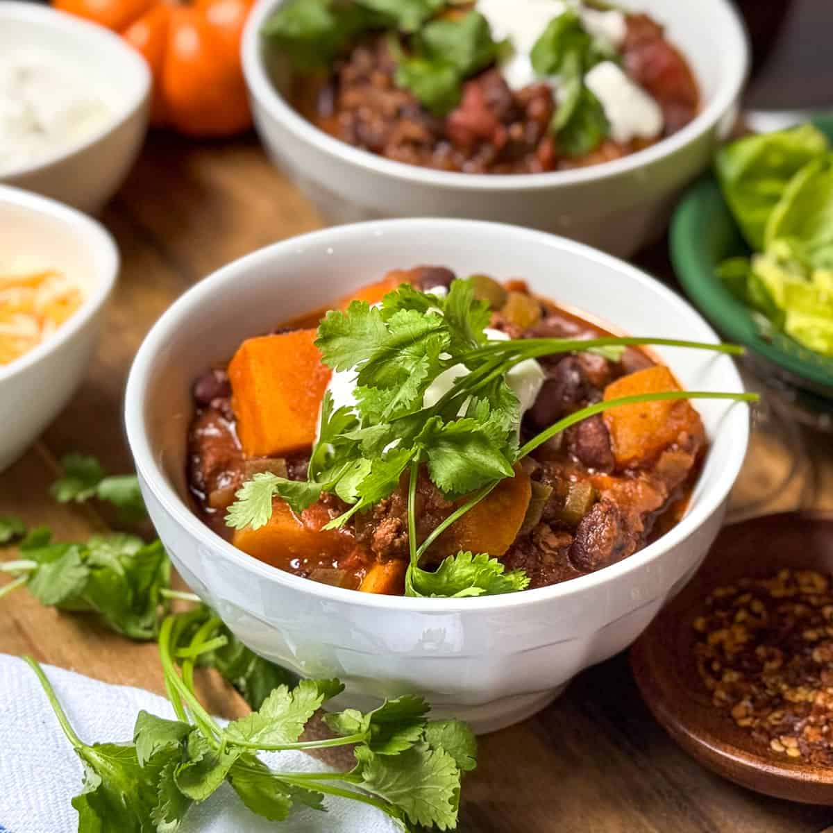 Bowl of Beef and Butternut Squash Chili, garnished with fresh cilantro, in a bowl with shredded cheese and sour cream on the side.