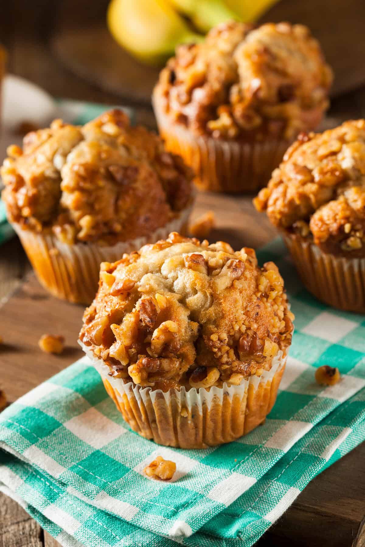 Vertical view of Banana Ginger Muffin sitting on a green and white kitchen linen with more in the background on a dark wooden surface.