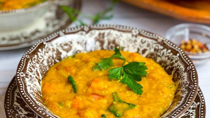 A bowl of Curry Lentil Soup with vegetables, garnished with parsley and served in a patterned bowl, accompanied by flatbread and salad in the background.