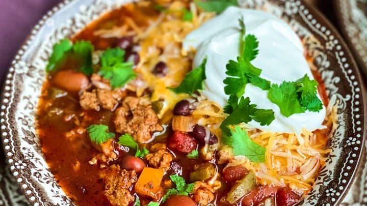 A bowl of Healthy Instant Pot Turkey Chili topped with shredded cheese, sour cream, and cilantro, served on a decorative plate with cornbread in the background.