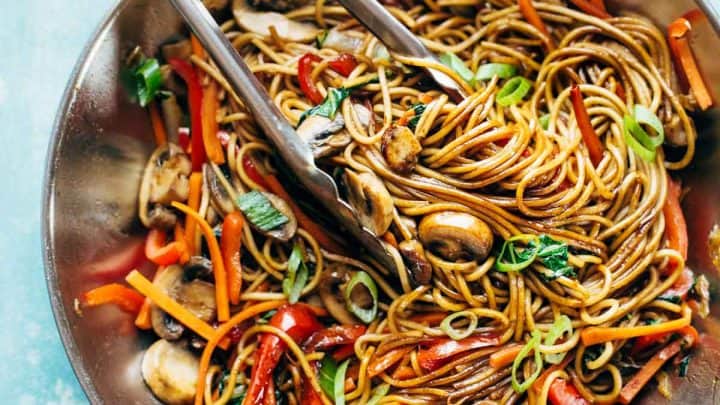 A hand uses tongs to mix vegetable lo mein—one of those fall family favorites—with mushrooms, bell peppers, carrots, and scallions in a stainless steel pan.