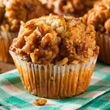 A close-up of a Banana Ginger Muffin in a paper liner, sitting on a green and white checkered cloth. Other muffins are blurred in the background.
